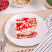 A slice of Heaven On Earth Cake, layered with cherries and cream and topped with sliced almonds, sits on a white plate beside a fork and striped napkin. Bowls of cherries and almonds complete the inviting scene.