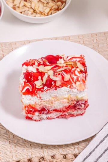 A slice of Heaven On Earth Cake, layered with cherries and cream and topped with sliced almonds, sits on a white plate beside a fork and striped napkin. Bowls of cherries and almonds complete the inviting scene.