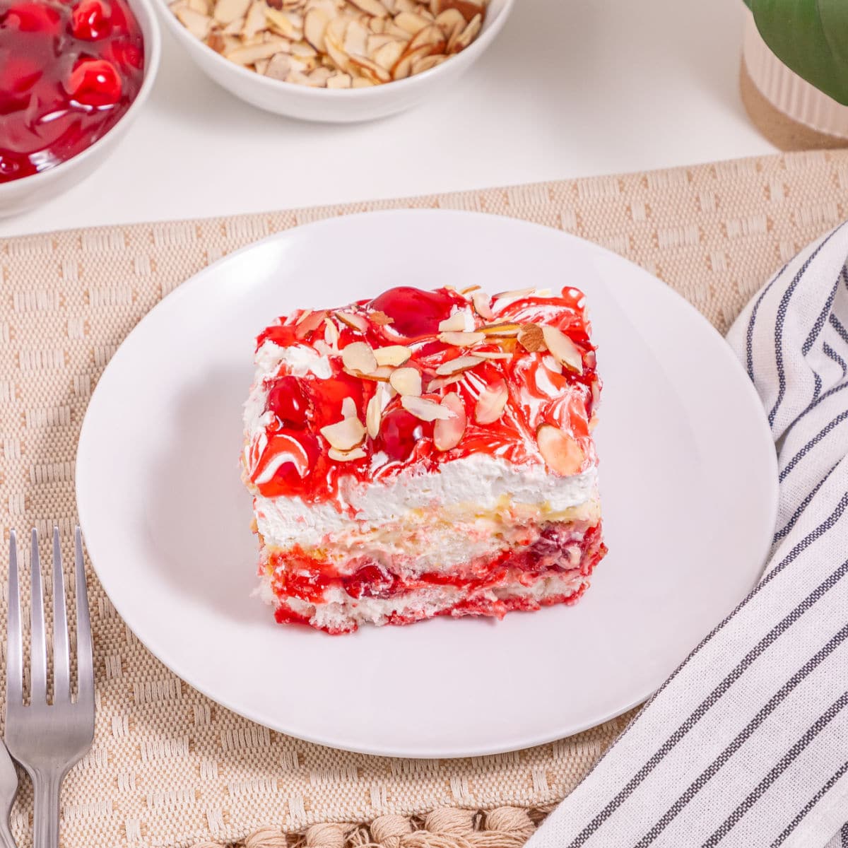 A slice of Heaven On Earth Cake, layered with cherries and cream and topped with sliced almonds, sits on a white plate beside a fork and striped napkin. Bowls of cherries and almonds complete the inviting scene.