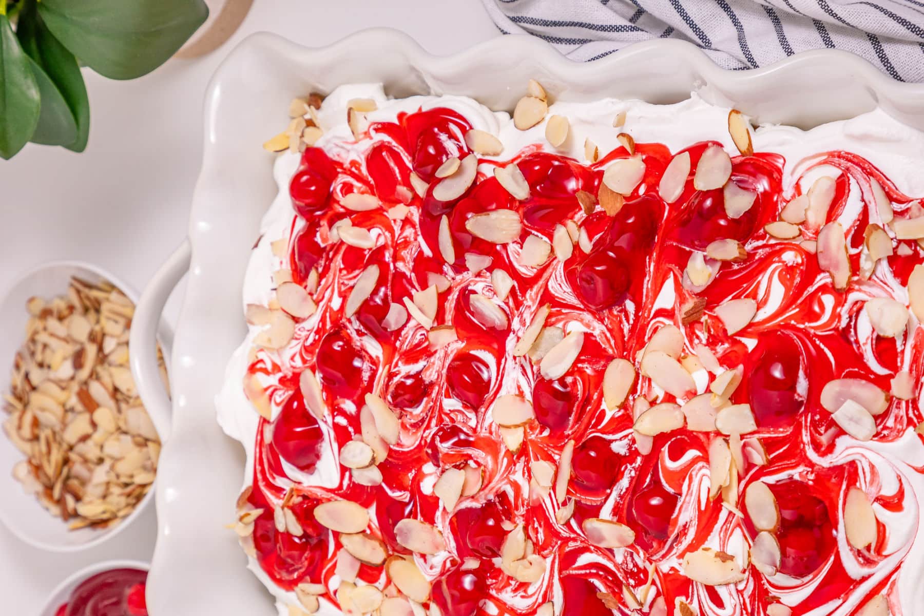 A square white dish filled with a dessert topped with swirled red cherry sauce, whipped cream, and sprinkled sliced almonds. Nearby are a bowl of sliced almonds, a plant, and a striped cloth.