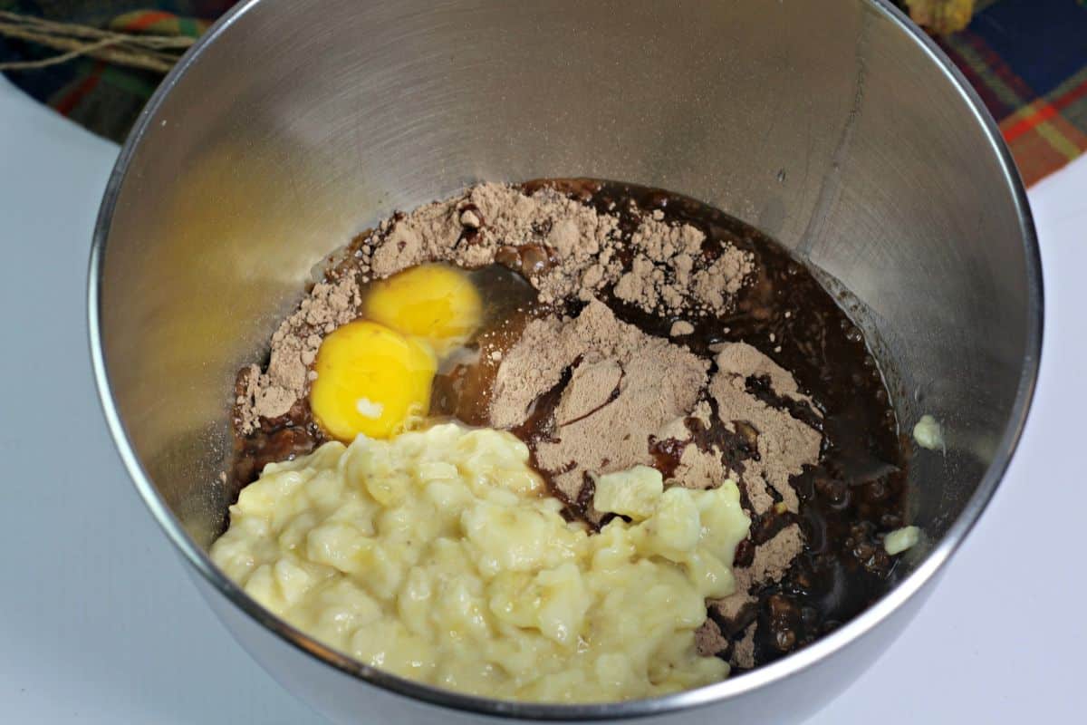 A metal mixing bowl containing two eggs, mashed banana, chocolate cake mix powder, and a dark liquid, ready to be mixed for baking.