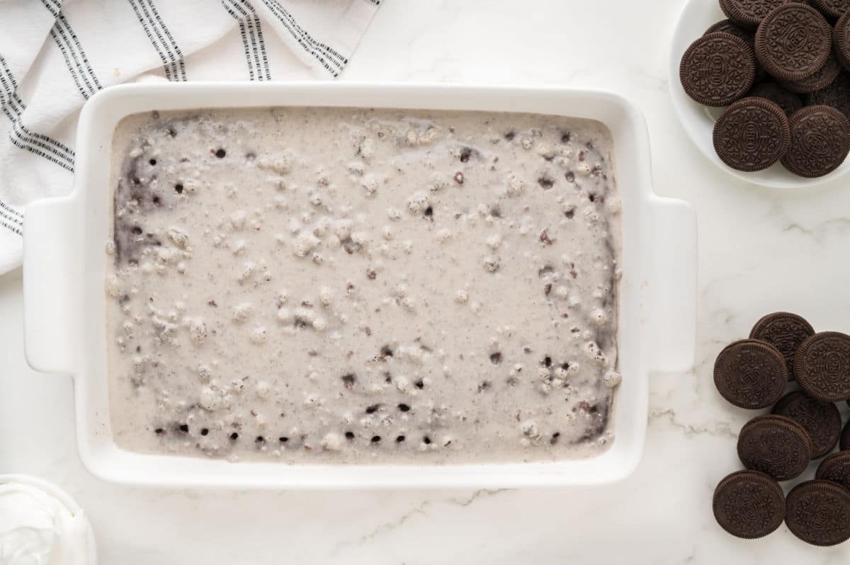 A white rectangular baking dish filled with an Oreo poke cake mixture sits on a marble surface. Whole chocolate sandwich cookies are nearby, along with a kitchen towel and a bowl of whipped topping.