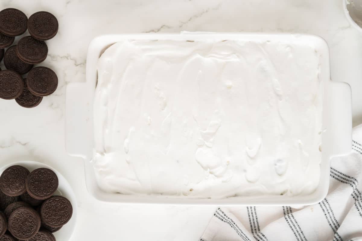 A white rectangular dish filled with a layer of fluffy whipped topping, surrounded by chocolate sandwich cookies on a white marble surface, showcases an irresistible Oreo poke cake, with a white and black striped cloth nearby.