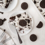 A plate with a piece of Oreo poke cake topped with whipped cream and cookie crumbles, garnished with a whole chocolate sandwich cookie. A fork, whole cookies, and a striped napkin are nearby on a white surface.