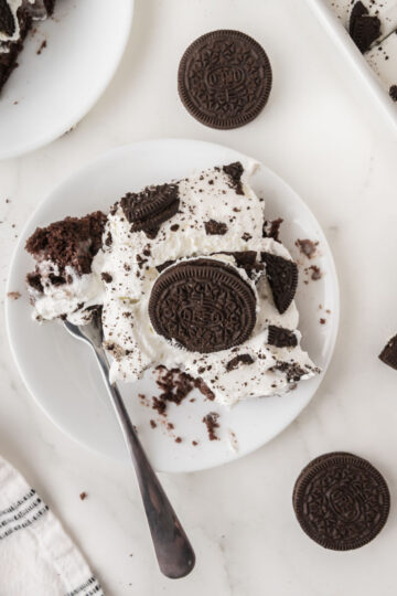 A plate with a piece of Oreo poke cake topped with whipped cream and cookie crumbles, garnished with a whole chocolate sandwich cookie. A fork, whole cookies, and a striped napkin are nearby on a white surface.