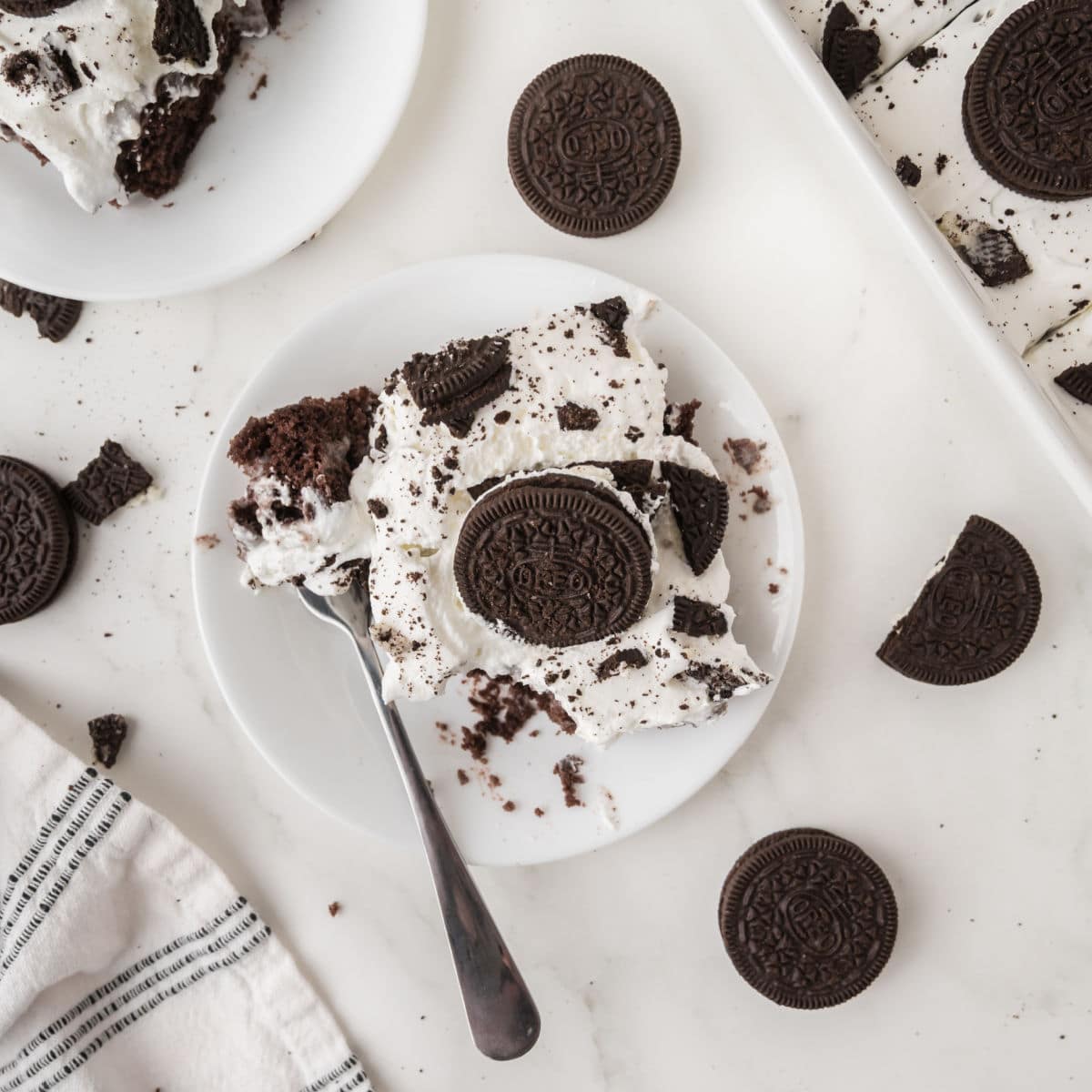 A plate with a piece of Oreo poke cake topped with whipped cream and cookie crumbles, garnished with a whole chocolate sandwich cookie. A fork, whole cookies, and a striped napkin are nearby on a white surface.