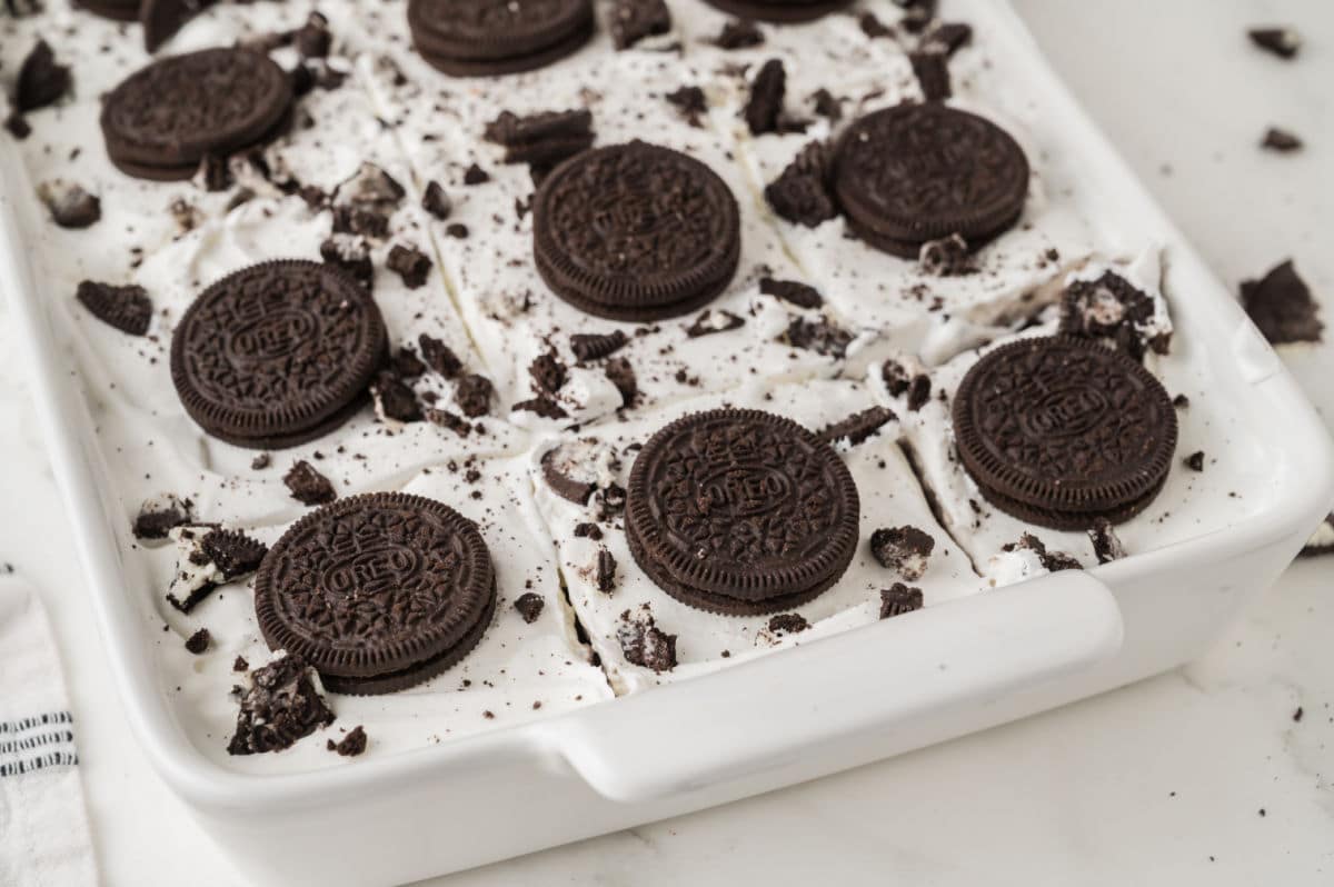 A white baking dish filled with oreo poke cake, topped with whole and crushed chocolate sandwich cookies over a layer of creamy white topping, cut into squares.