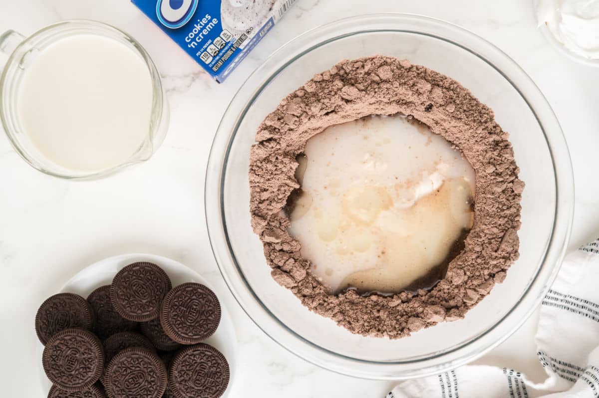 A mixing bowl with chocolate cake mix, milk, and other wet ingredients for an Oreo poke cake sits in the center. Nearby are a measuring cup of milk, a plate of sandwich cookies, a striped towel, and a box of Oreo cookies on a white countertop.