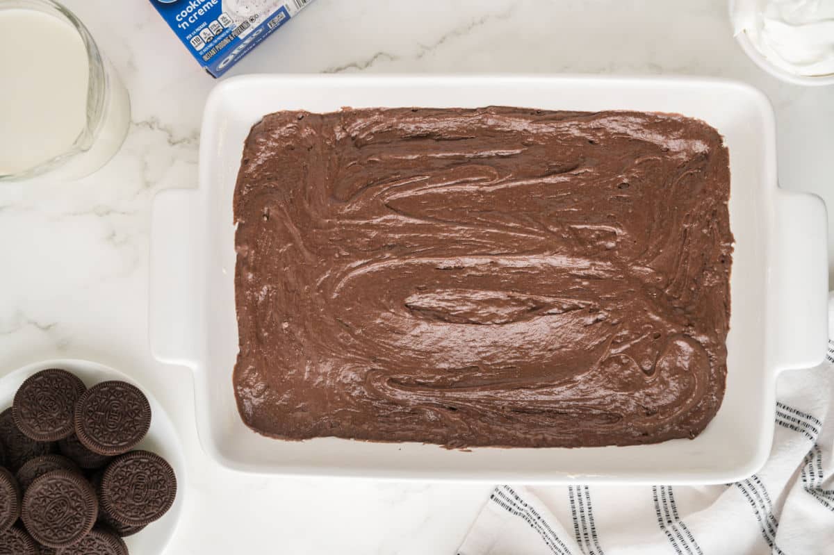 A white baking dish filled with chocolate batter sits on a marble surface, ready to become a decadent Oreo poke cake. Nearby are a glass of milk, chocolate sandwich cookies, whipped topping, and a striped kitchen towel.