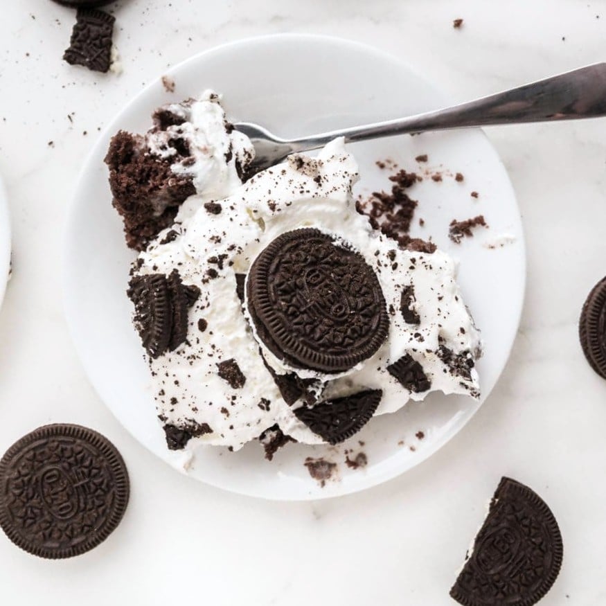 A dessert with chocolate cookies and whipped cream is served on a white plate with a spoon. Several whole cookies and crumbs are scattered around, evoking the indulgent look of an Oreo poke cake on a white surface.