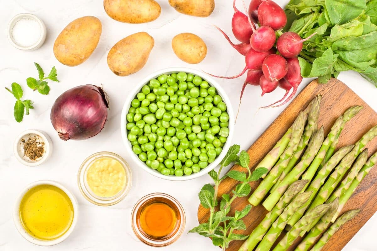 A variety of fresh vegetables and ingredients, including potatoes, radishes, asparagus, red onion, green peas, olive oil, mustard, vinegar, herbs, and seasonings, arranged on a white surface and wooden cutting board.