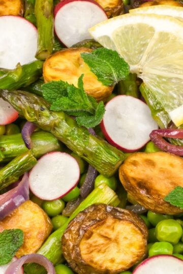 Colorful salad with roasted sweet potato slices, asparagus, green peas, radish slices, red onion, fresh mint leaves, and lemon wedges on top. The ingredients are fresh and vividly arranged.
