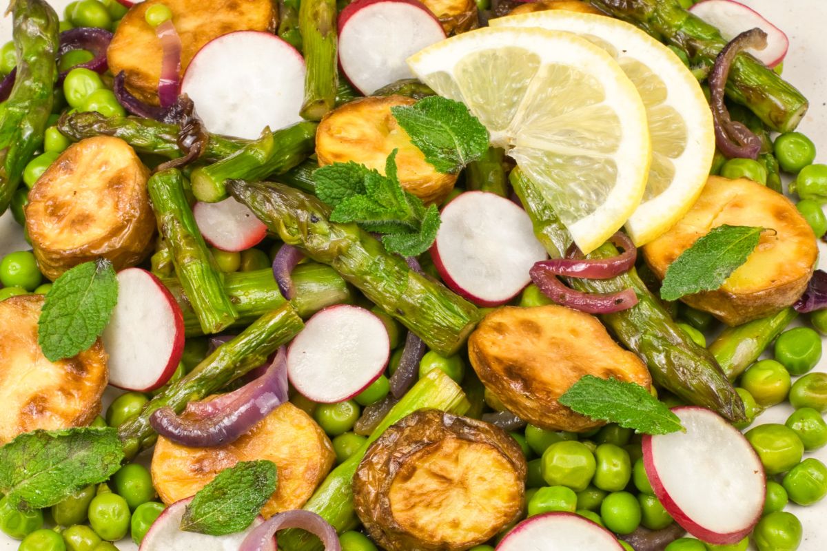 Colorful salad with roasted sweet potato slices, asparagus, green peas, radish slices, red onion, fresh mint leaves, and lemon wedges on top. The ingredients are fresh and vividly arranged.