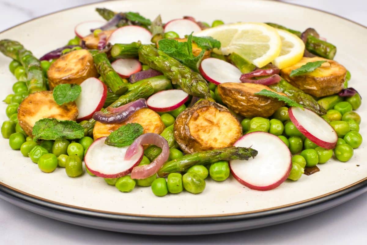 A plate of salad with roasted potatoes, asparagus, green peas, radish slices, red onion, mint leaves, and lemon slices arranged on top.
