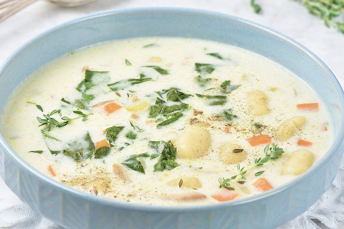 A bowl of creamy chicken gnocchi soup with spinach, carrots, and herbs, garnished with fresh thyme and black pepper. The soup is served in a light blue bowl on a light surface.