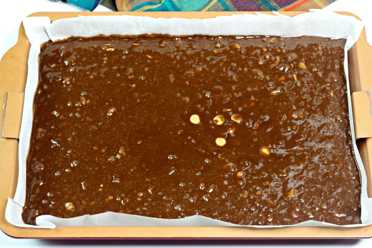 A rectangular baking pan lined with parchment paper is filled with unbaked chocolate brownie batter containing visible white peanut butter chips.