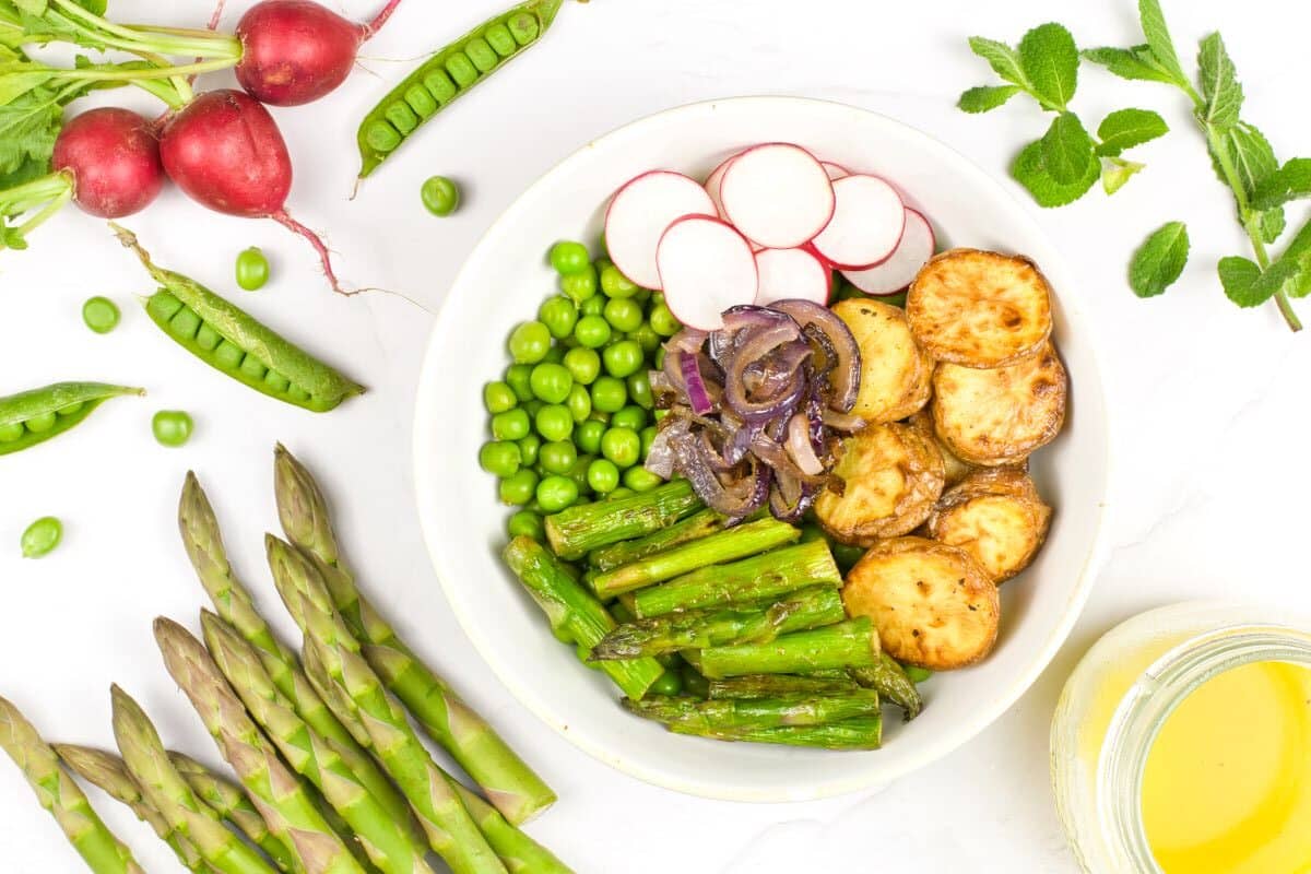 A bowl filled with roasted potatoes, green peas, asparagus, sliced radishes, and caramelized onions. Surrounding the bowl are fresh asparagus, radishes, pea pods, mint leaves, and a small bowl of yellow sauce.