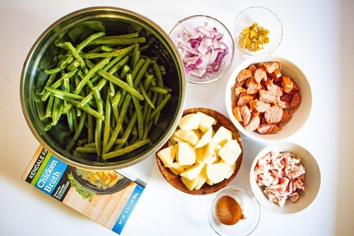 A top-down view of bowls containing fresh green beans, diced red onions, chopped yellow peppers, sliced sausage, cubed potatoes, chopped bacon, and a seasoning blend, with a box of chicken broth on the side.