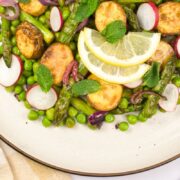 A plate of roasted potatoes, asparagus, peas, radish slices, and red onions, garnished with mint leaves and lemon slices. A beige napkin and spoon are partially visible beside the plate.