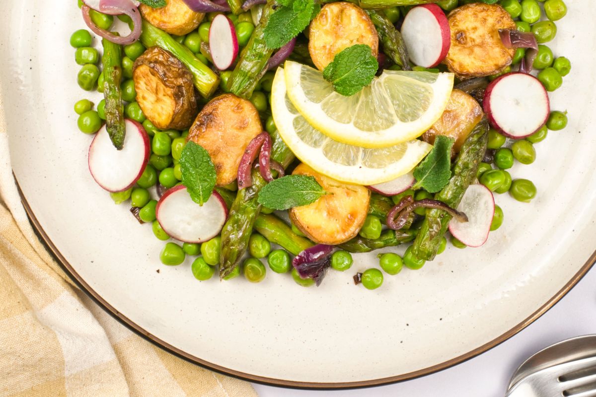 A plate of roasted potatoes, asparagus, peas, radish slices, and red onions, garnished with mint leaves and lemon slices. A beige napkin and spoon are partially visible beside the plate.