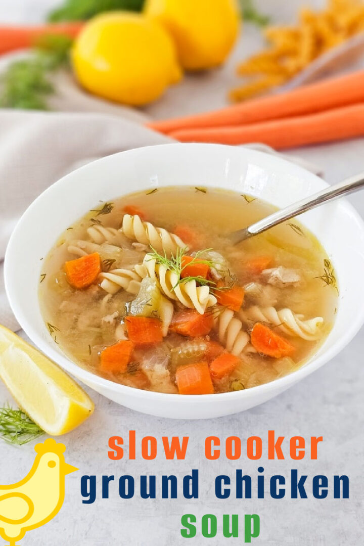 A white bowl of ground chicken soup with rotini pasta, carrots, and celery sits on a table with a spoon inside. Fresh carrots, lemons, dill, and a lemon wedge surround the bowl. Text reads: slow cooker ground chicken soup.