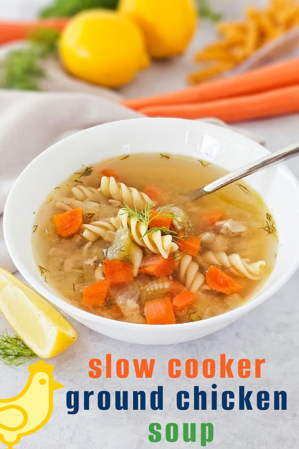 A white bowl of ground chicken soup with rotini pasta, carrots, and celery sits on a table with a spoon inside. Fresh carrots, lemons, dill, and a lemon wedge surround the bowl. Text reads: slow cooker ground chicken soup.