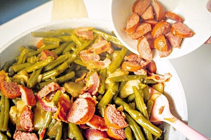 Sliced sausage being added to a pan filled with cooked green beans and chopped onions, with a pink spoon visible in the mixture.