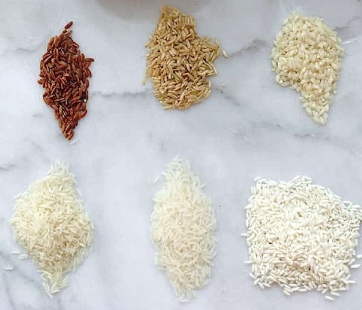 Six different varieties of uncooked rice arranged in two rows on a white marble surface, each pile displaying distinct colors and shapes.