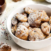A white bowl filled with small, round Air Fryer Funnel Cake Bites dusted with powdered sugar and colorful sprinkles sits on a white surface, with more bites and sprinkles scattered nearby.