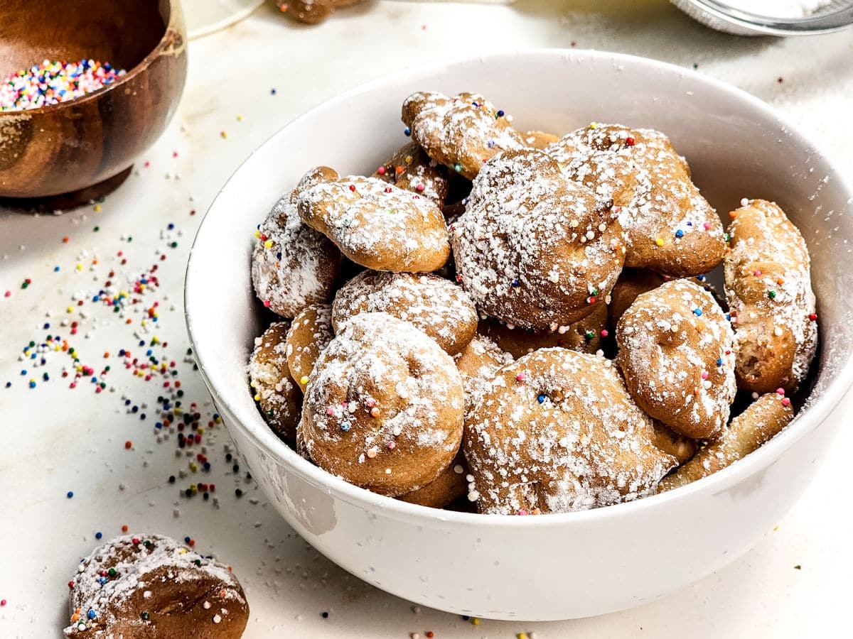 A white bowl filled with small, round Air Fryer Funnel Cake Bites dusted with powdered sugar and colorful sprinkles sits on a white surface, with more bites and sprinkles scattered nearby.