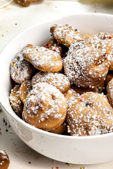A white bowl filled with round, golden Air Fryer Funnel Cake Bites topped with powdered sugar and colorful sprinkles. Some cake bites and sprinkles are scattered on the white surface around the bowl.