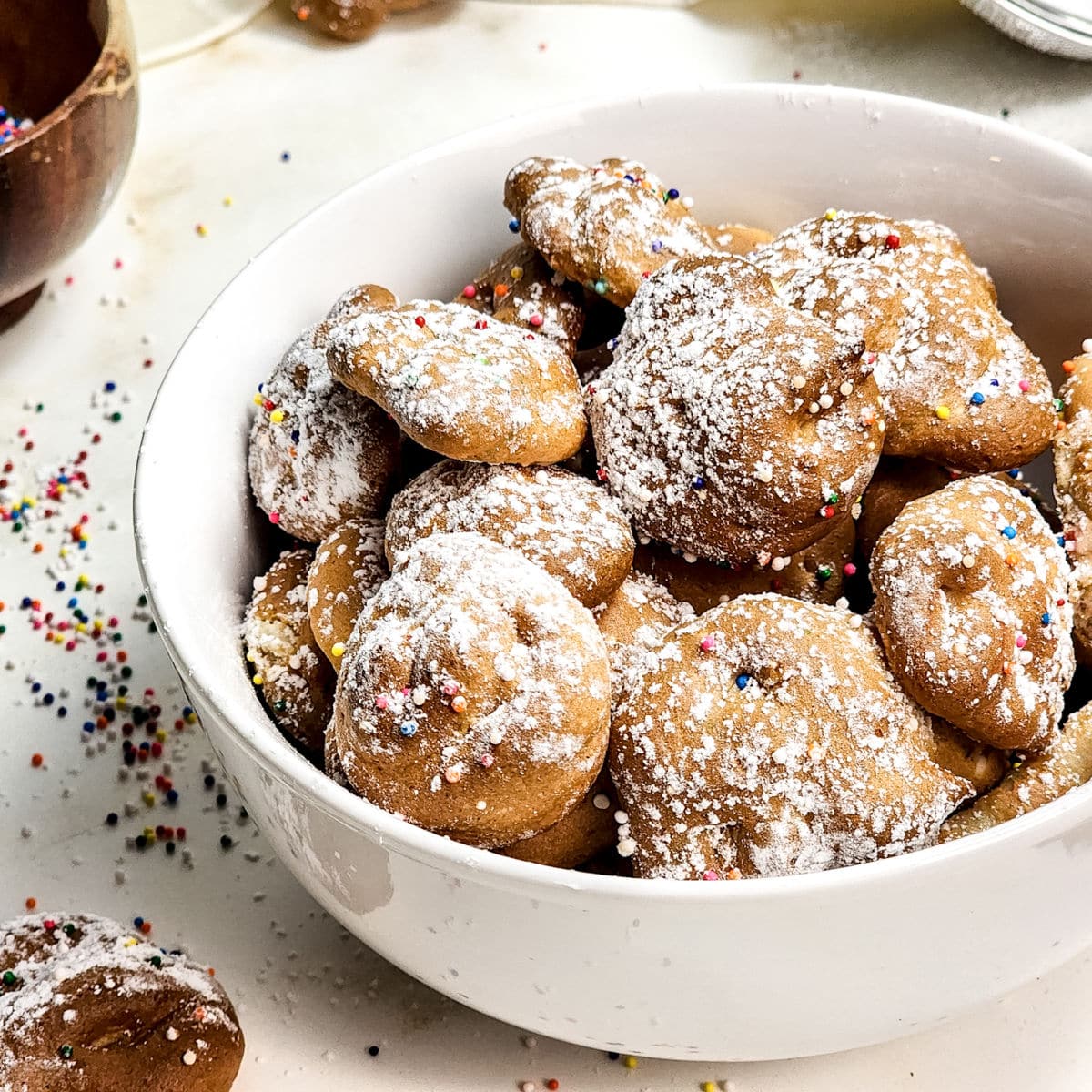 A white bowl filled with round, golden Air Fryer Funnel Cake Bites topped with powdered sugar and colorful sprinkles. Some cake bites and sprinkles are scattered on the white surface around the bowl.