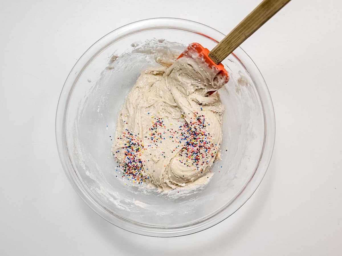 A glass bowl filled with cookie dough and colorful sprinkles, being mixed with a red spatula on a white surface—perfect for whipping up fun treats like Air Fryer Funnel Cake Bites.
