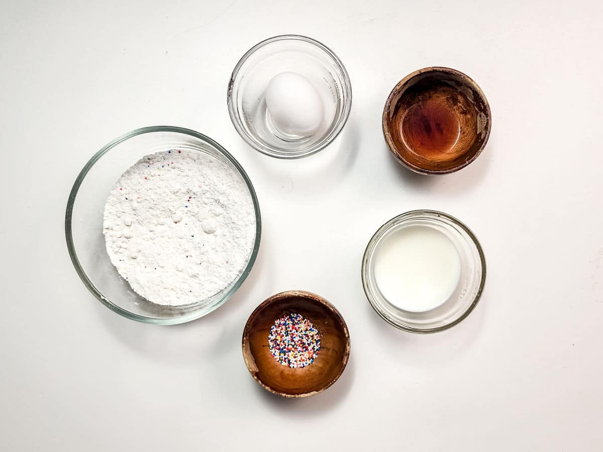 Five ingredients for Air Fryer Funnel Cake Bites are arranged on a white surface: a bowl with white powder, a small glass bowl with an egg, a wooden bowl with liquid, a jar of milk, and a wooden bowl filled with colorful sprinkles.