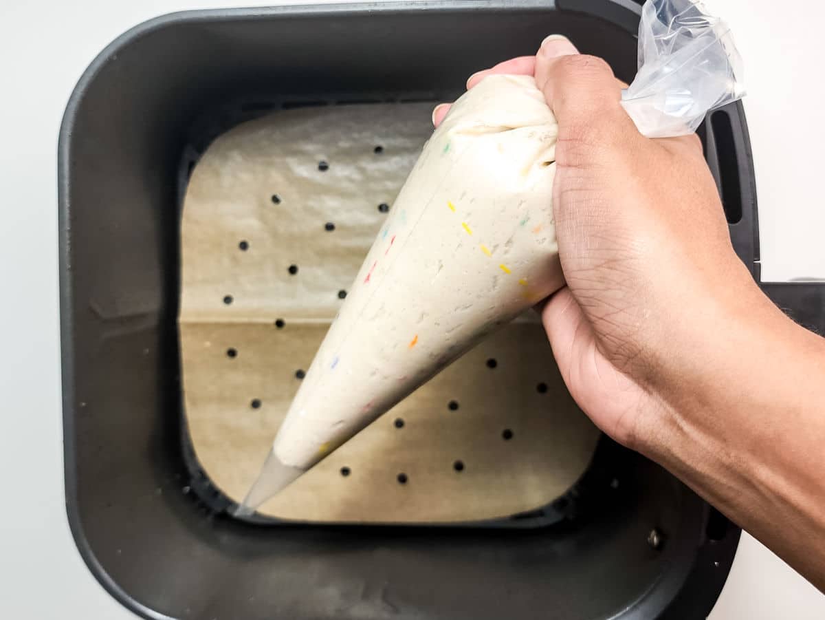 A hand holds a piping bag filled with cake batter over a parchment-lined air fryer basket, preparing to pipe Air Fryer Funnel Cake Bites onto the surface.