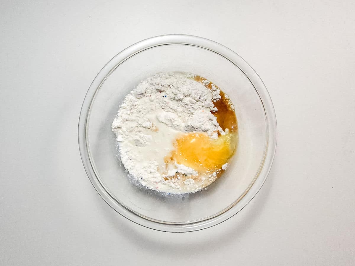 A glass mixing bowl containing flour, an egg, milk, and a liquid ingredient, set on a white surface before mixing for delicious Air Fryer Funnel Cake Bites.