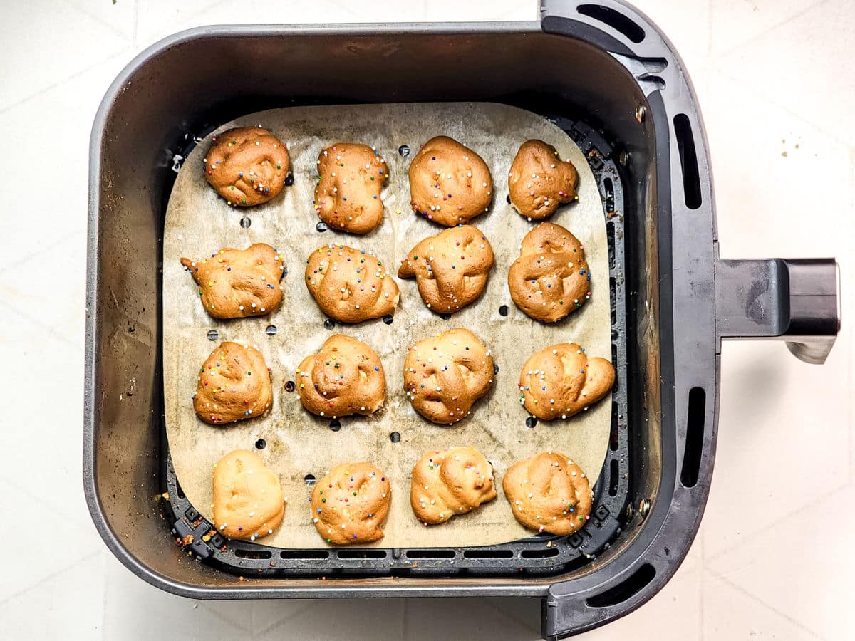 Round, golden-brown Air Fryer Funnel Cake Bites sit evenly spaced on parchment paper inside an air fryer basket, topped with colorful sprinkles. The background is a light, patterned surface.