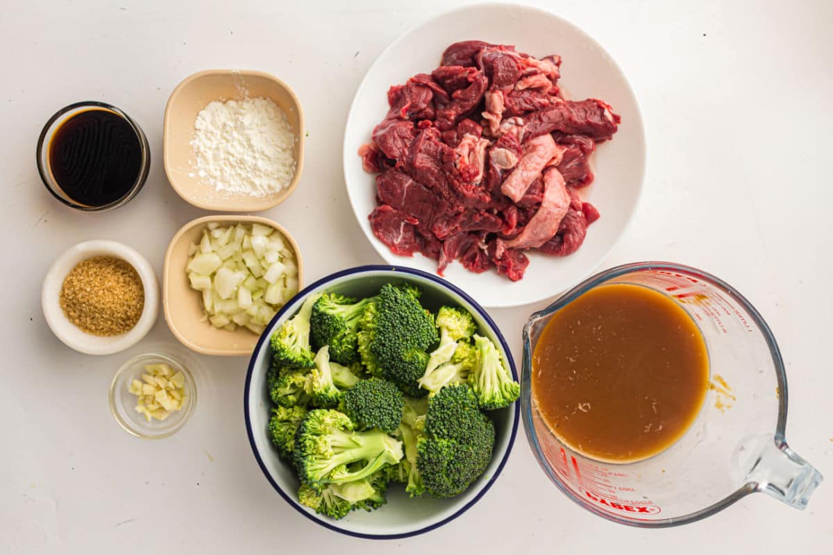 Various ingredients for a beef and broccoli slow cooker dish are arranged on a white surface, including sliced raw beef, chopped broccoli, diced onion, minced garlic, brown sauce, soy sauce, cornstarch, and brown sugar.