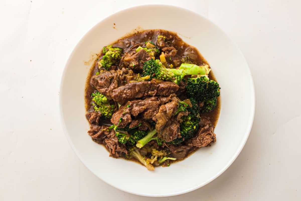 A white plate filled with beef and broccoli in a brown sauce, likely a stir-fry dish, placed on a white surface—perfect for recreating an easy beef and broccoli slow cooker meal at home.