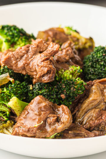 A close-up of a white bowl filled with beef and broccoli slow cooker stir-fry, featuring tender slices of beef and green broccoli florets coated in a savory brown sauce.