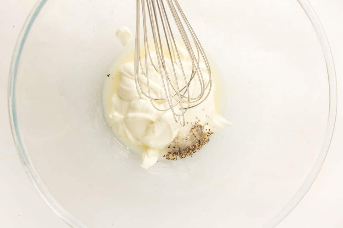 A metal whisk in a glass bowl containing a mixture of mayonnaise, black pepper, and a splash of milk, ready to be blended for a creamy chicken pasta with bacon and peas.