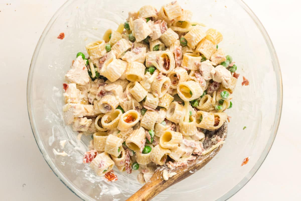 A glass bowl filled with creamy chicken pasta with bacon and peas, featuring tube-shaped pasta, sun-dried tomatoes, and tender chicken pieces, all mixed together with a wooden spoon resting inside the bowl.