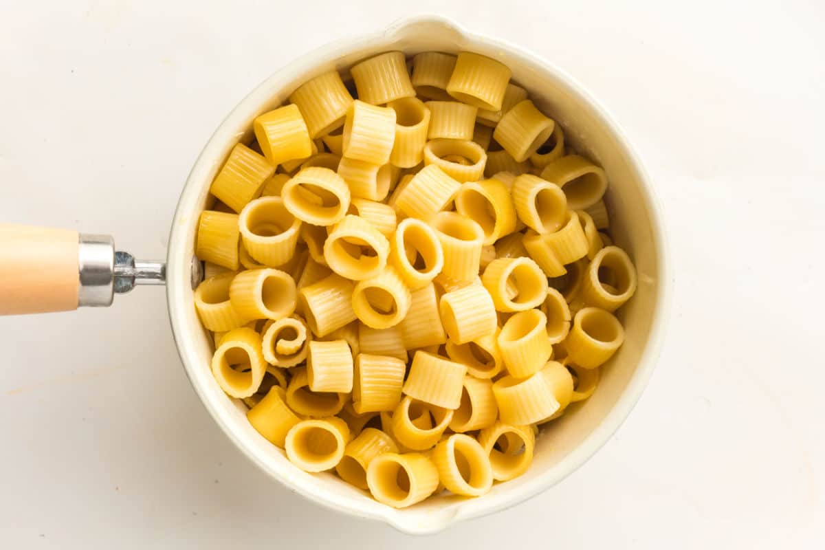 A white pot filled with cooked ditalini pasta for chicken pasta with bacon and peas sits on a white surface, with a wooden-handled utensil visible on the left side.