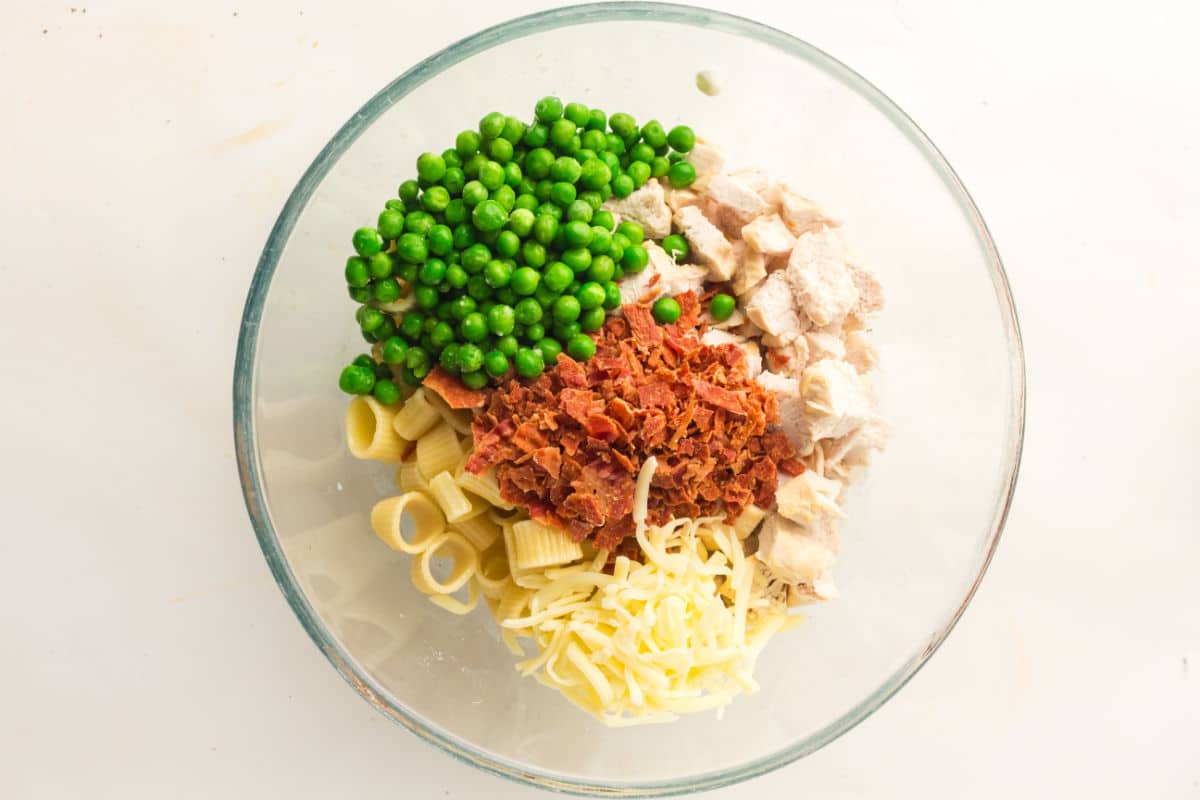 A glass bowl containing uncooked chicken pasta with bacon and peas, along with shredded cheese and cooked chicken pieces, all arranged in separate sections.