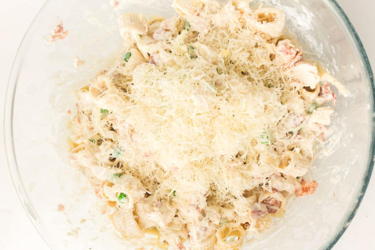 A glass bowl filled with creamy chicken pasta with bacon and peas, topped with a generous layer of grated cheese for extra flavor.