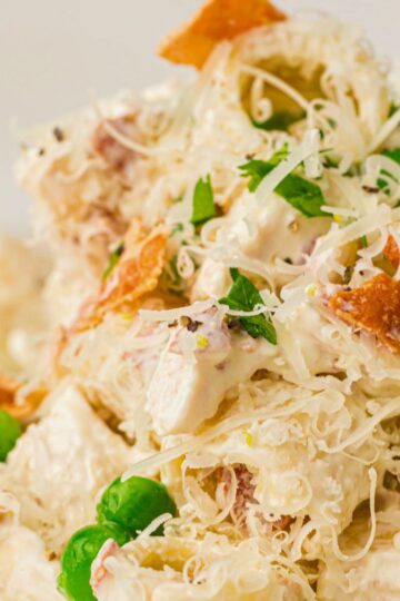 A close-up of a creamy chicken pasta with bacon and peas, featuring shredded cheese, green peas, chopped parsley, and chunks of chicken—all mixed together and finished with grated cheese and herbs.