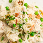 Close-up of a creamy chicken pasta with bacon and peas, topped with shredded cheese, chopped herbs, diced ham, and small pasta pieces on a white plate.