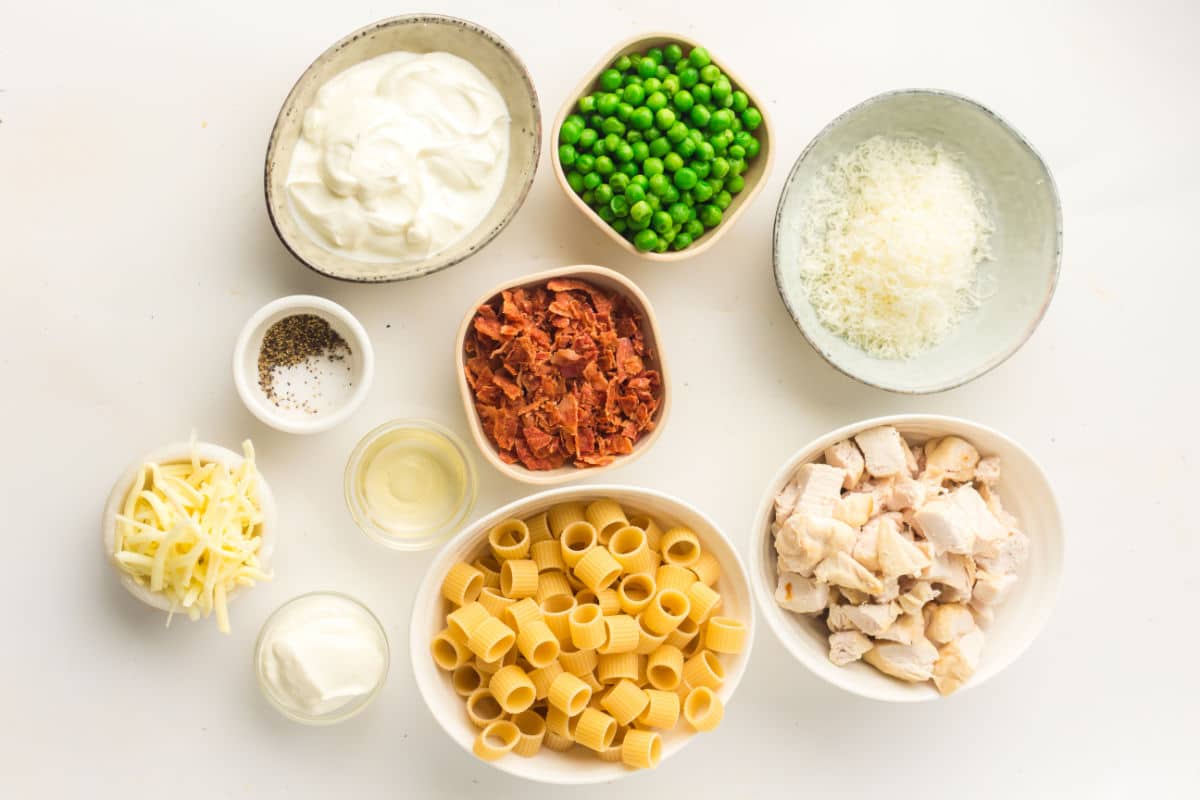 Bowls of ingredients on a white surface, including rigatoni pasta, cooked chicken, shredded cheese, green peas, grated cheese, sour cream, sun-dried tomatoes, ground pepper, oil—a perfect spread for chicken pasta with bacon and peas.