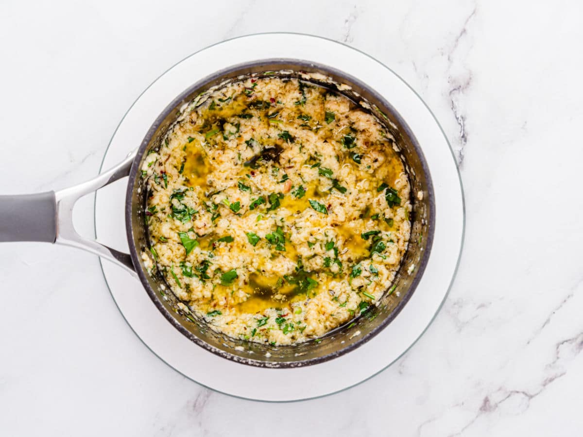 A pot filled with melted butter mixed with chopped herbs and minced garlic, perfect for drizzling over Garlic Parmesan Chicken Air Fryer recipes, sits on a white plate atop a marble countertop.