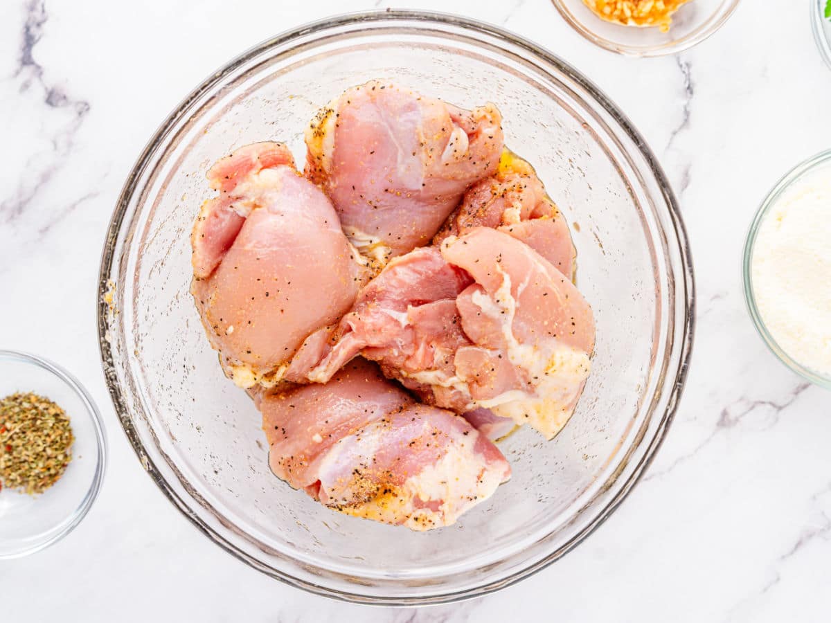 Raw, seasoned chicken thighs in a glass bowl on a white marble surface, surrounded by small bowls containing spices, garlic, and breadcrumbs—perfect for making Garlic Parmesan Chicken Air Fryer style.